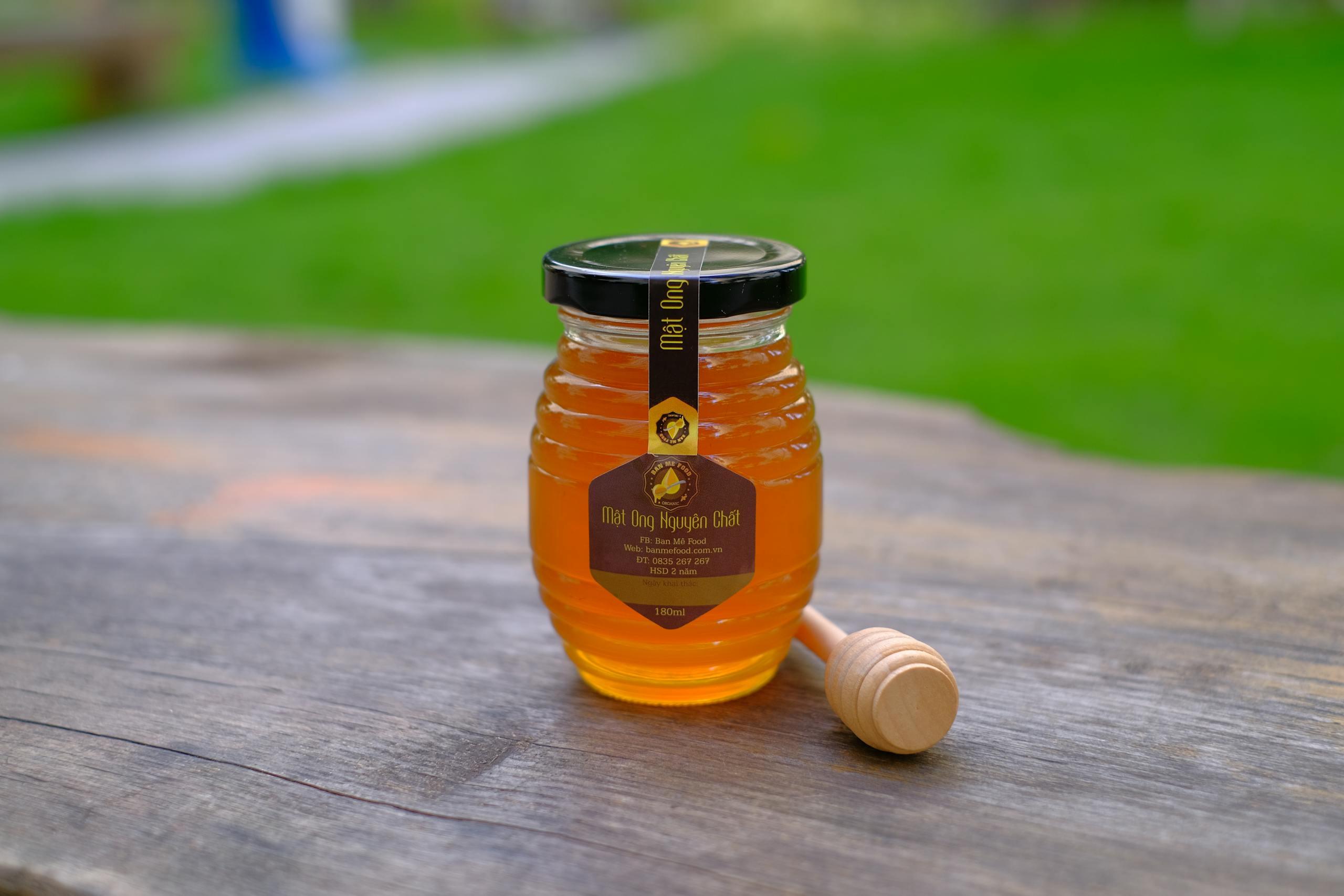 A glass jar of honey with a dipper on a rustic wooden table outdoors.