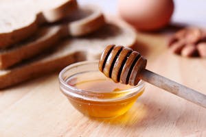 Close-up of honey dipper in a bowl with honey on wooden table beside sliced bread.