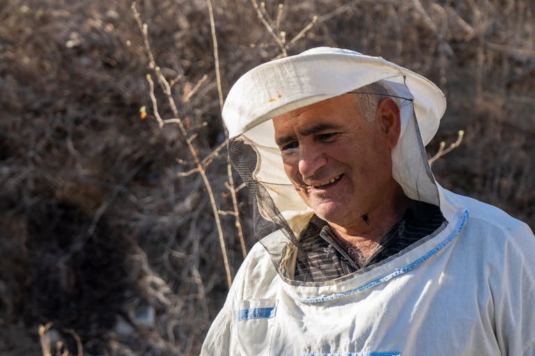 Senior beekeeper smiling outdoors in protective clothing during a sunny day.