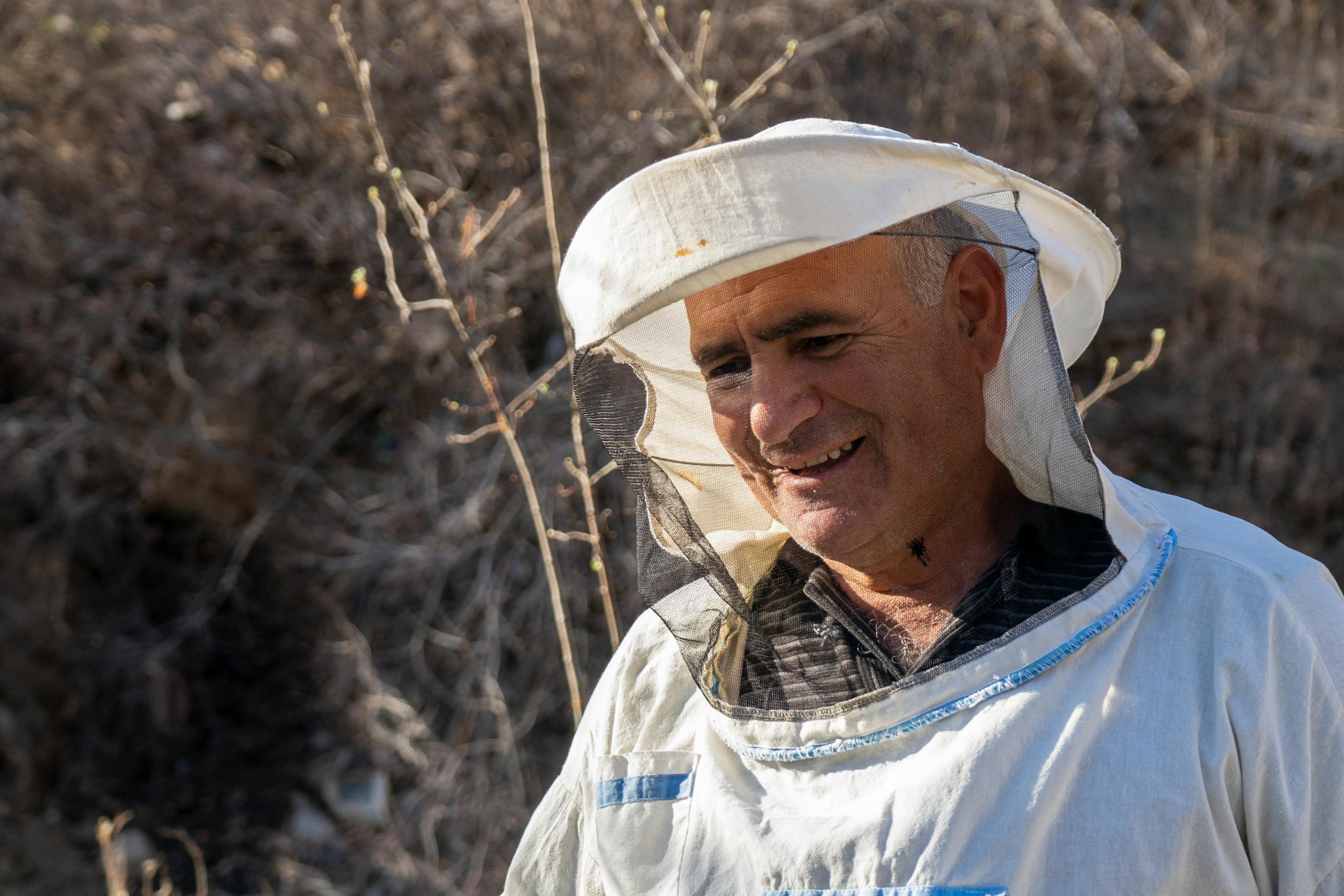 Senior beekeeper smiling outdoors in protective clothing during a sunny day.